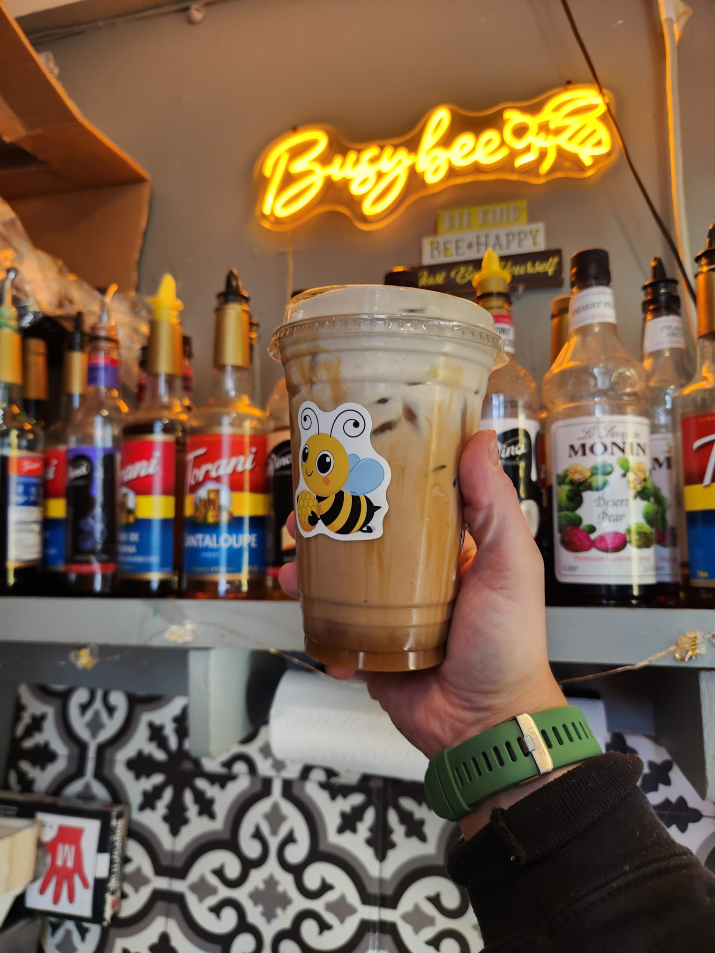 Iced latte in a Busy Bee Espresso cup held up beneath the neon shop sign